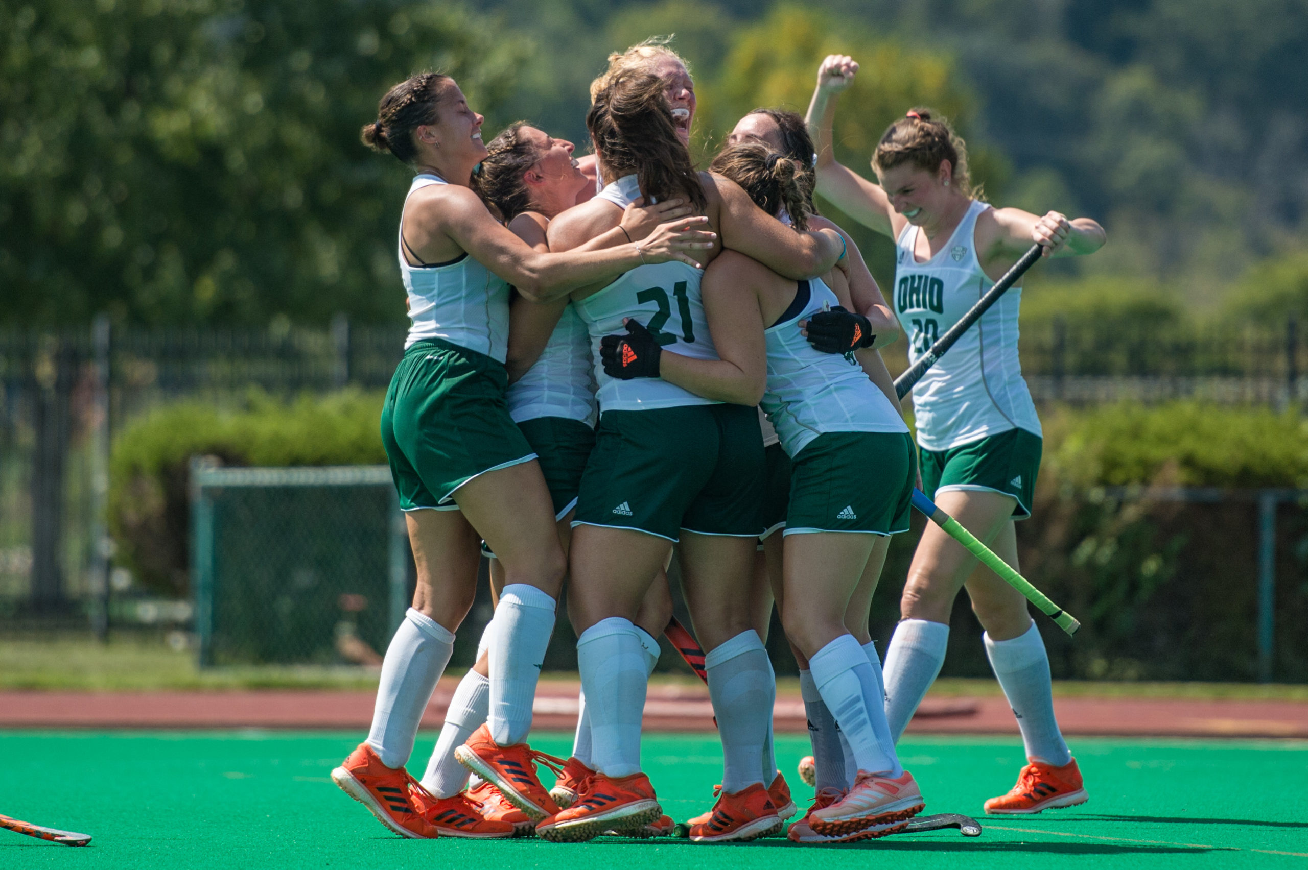 The Ohio University field hockey team celebrates in a group hug in white uniform tops and green kilts.