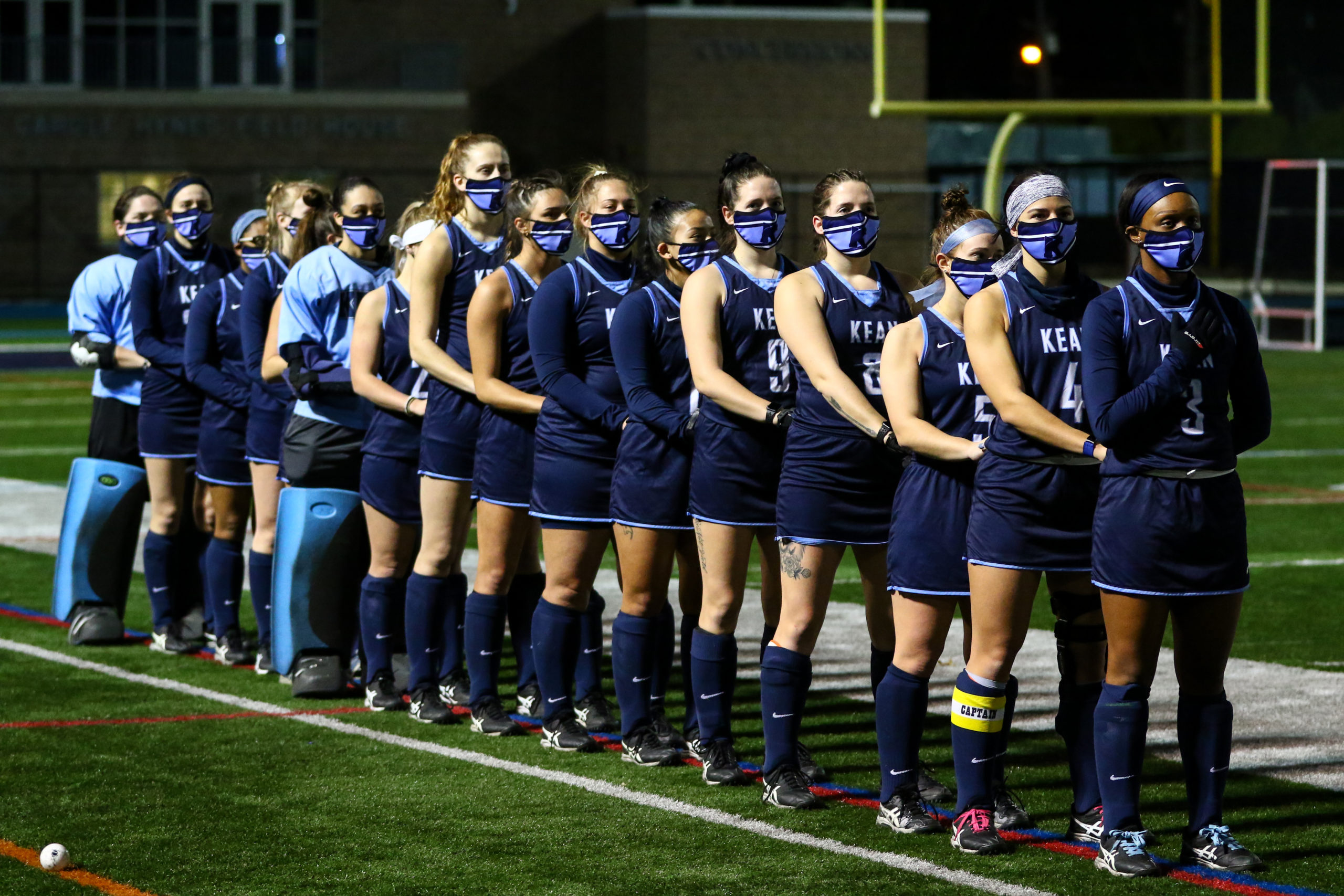 The Kean University field hockey team lined up at at the start of a game in dark blue uniforms and dark blue face masks.