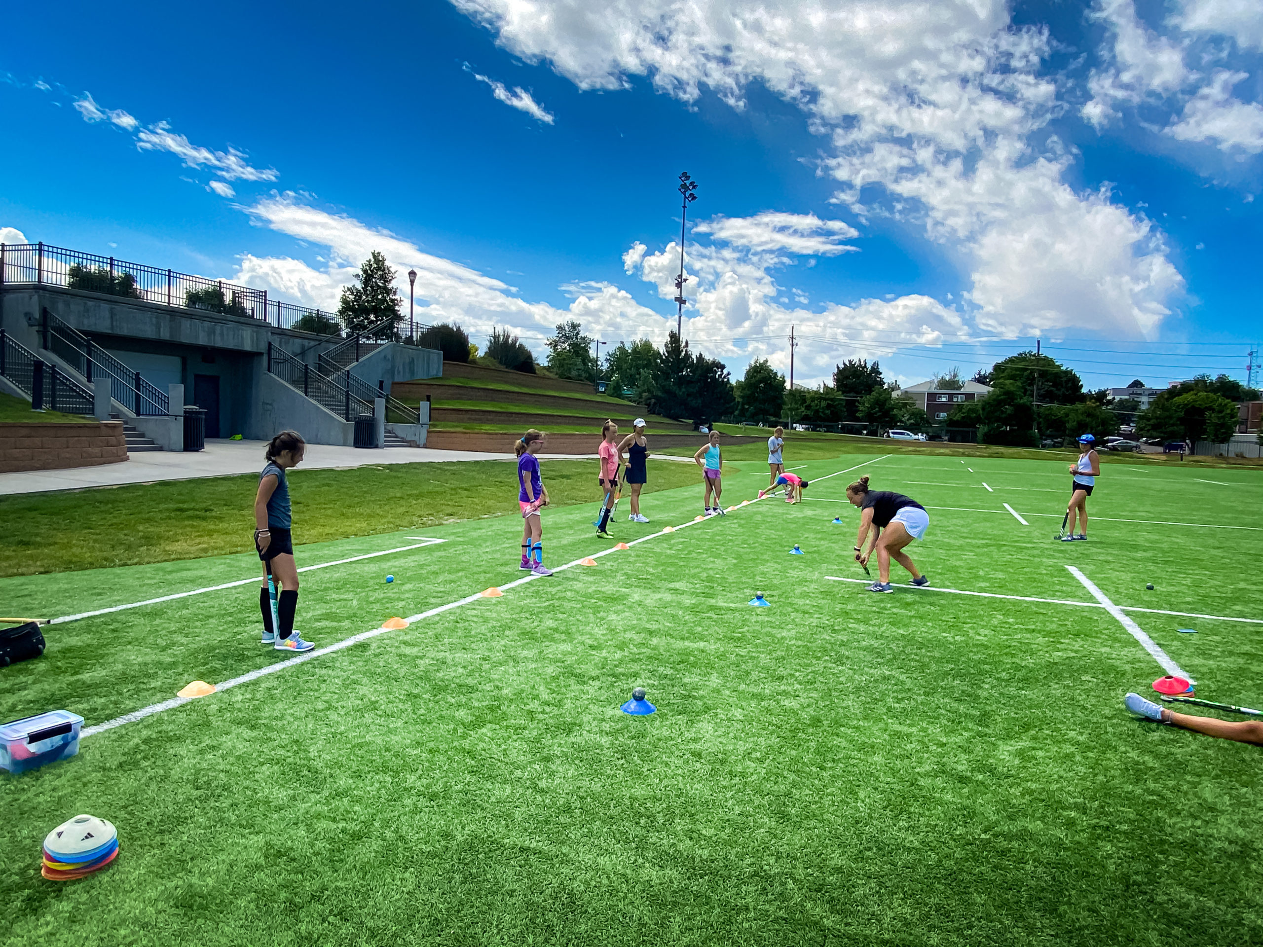 Sydney Supica demonstrates a skill to a group of young field hockey players on a green turf under blue sky and dramatic cumulous clouds.