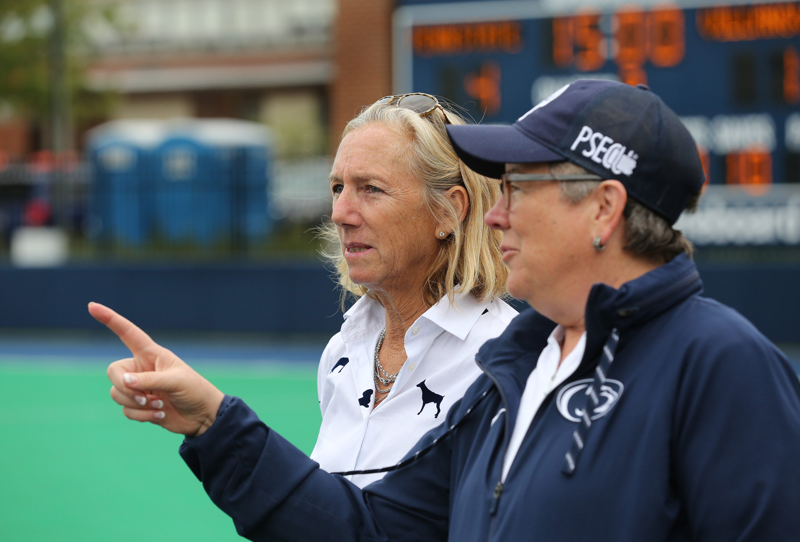 Charlene Morett-Curtiss, in a white pullover, and Sandy Barbour, in a navy blue pullover and hat, stand on the sidelines of a field hockey game.